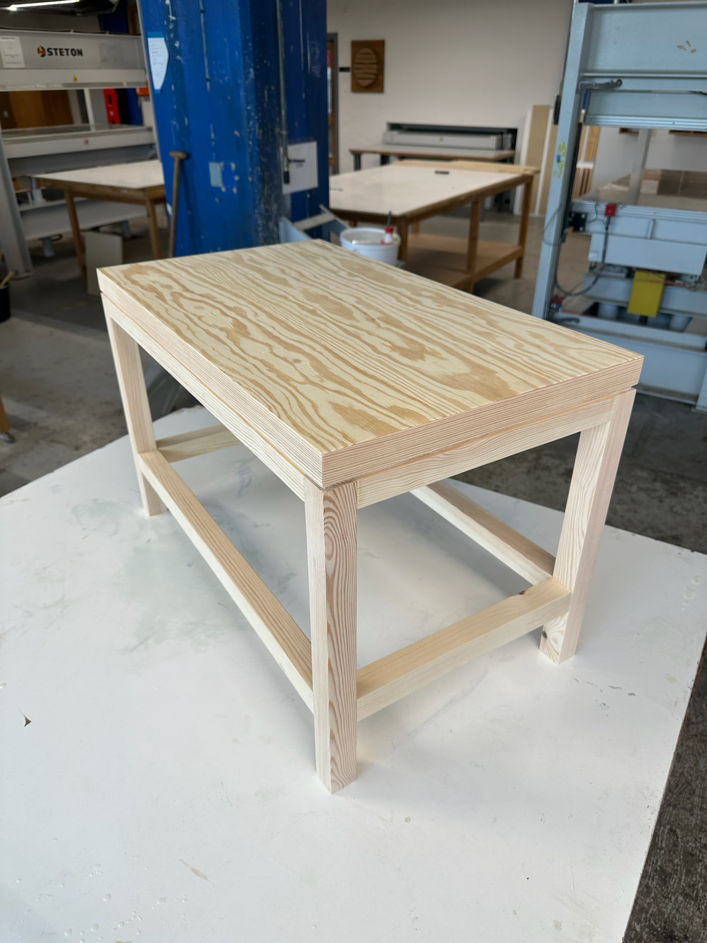 Finished pine bench with a plywood top, sitting on a white table in the workshop.