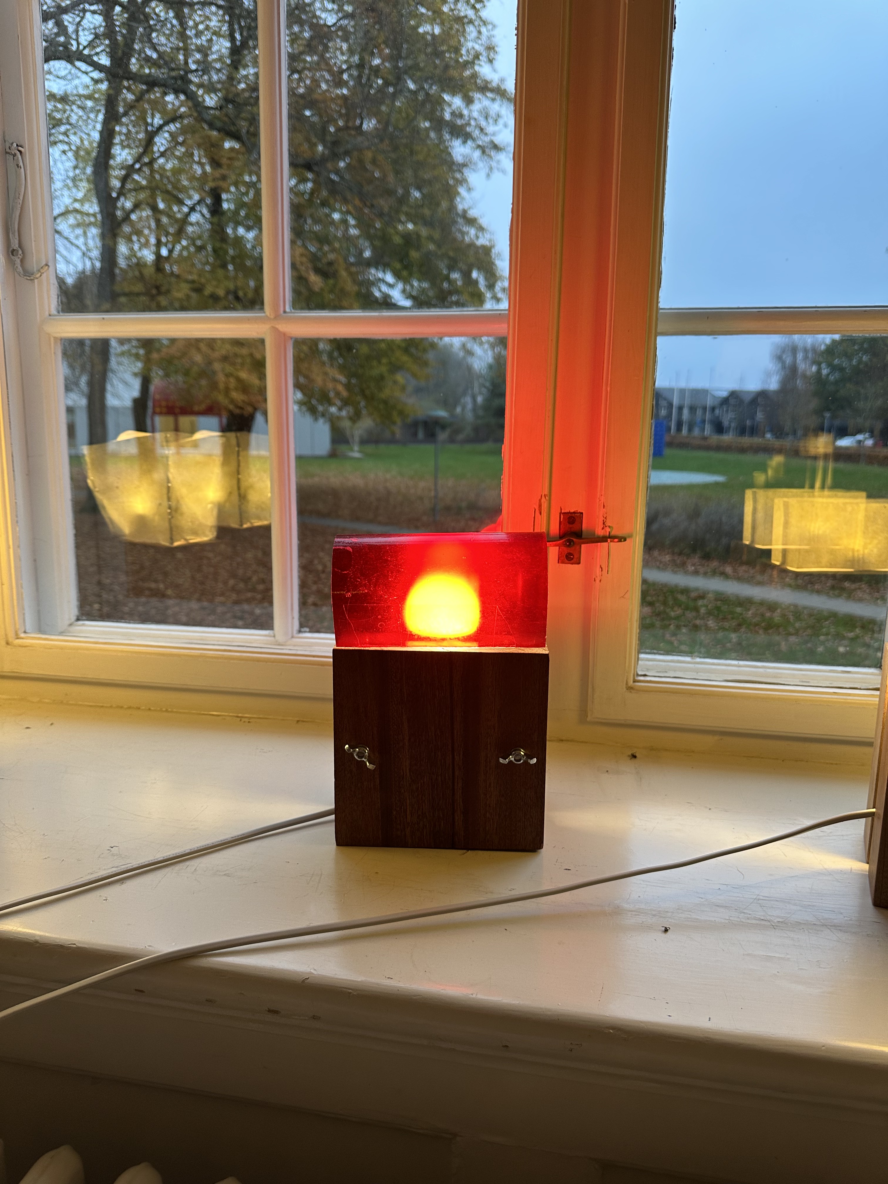 Bioplastic lamp with a red shade glowing on a windowsill at dusk.