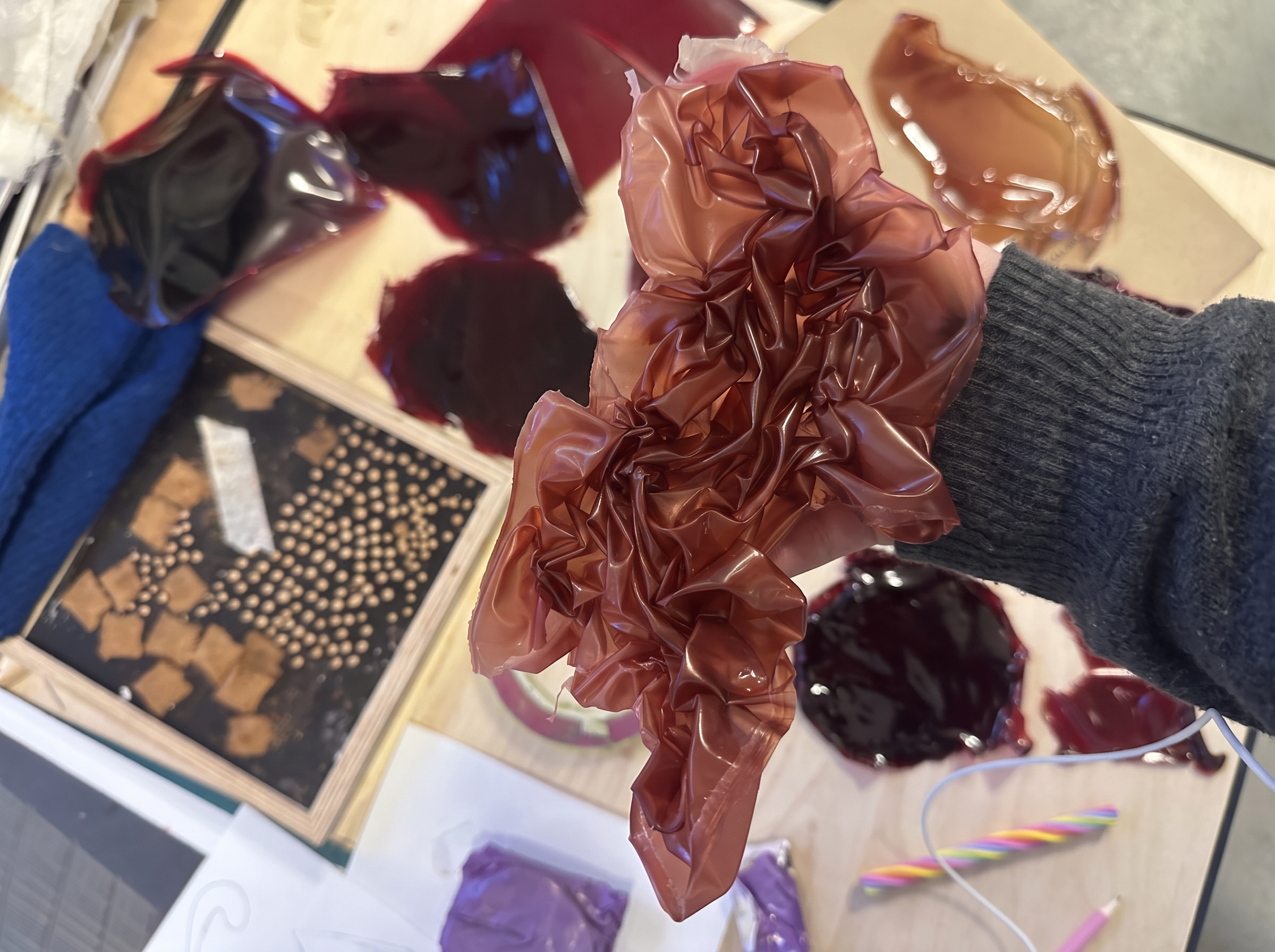 Hand holding a crumpled sheet of red-brown bioplastic among material samples on a worktable.